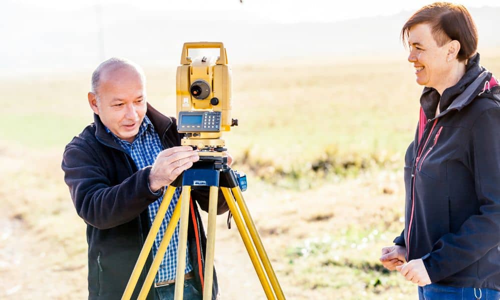 Licensed land surveyor performing an elevation survey at a residential property while the homeowner observes the process
