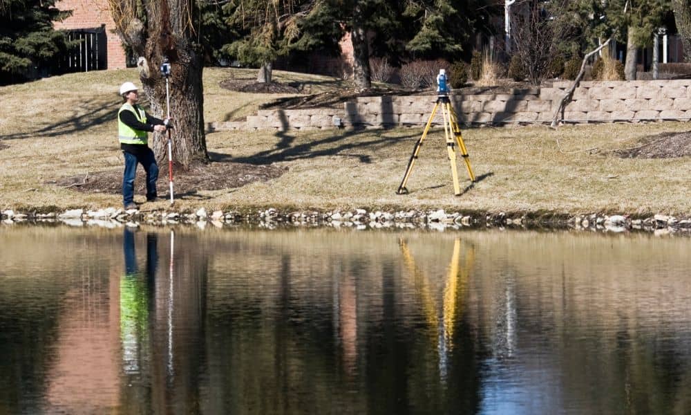 Land surveyor measuring home elevation near water - ALTA SURVEY Maine A licensed land surveyor measuring ground height to prepare an elevation certificate for a residential property