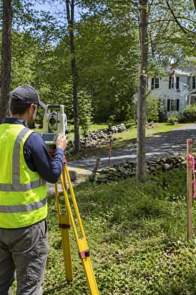 Land surveyor using a tripod-mounted instrument to measure property lines on-site, capturing survey mapping in real conditions